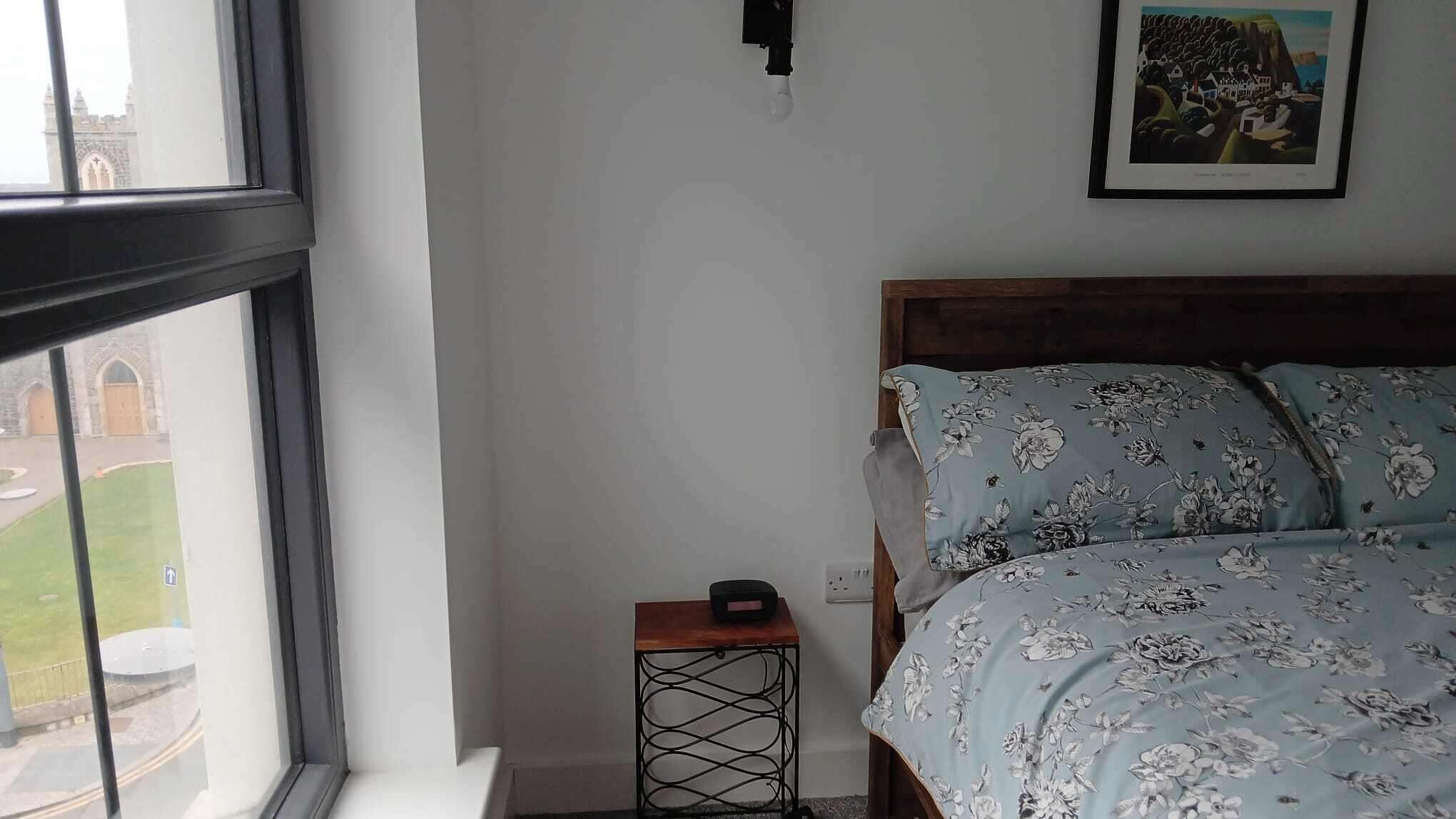 Une chambre à coucher comprenant un lit, une petite table, une lampe fixée au mur et une fenêtre donnant sur un immeuble.