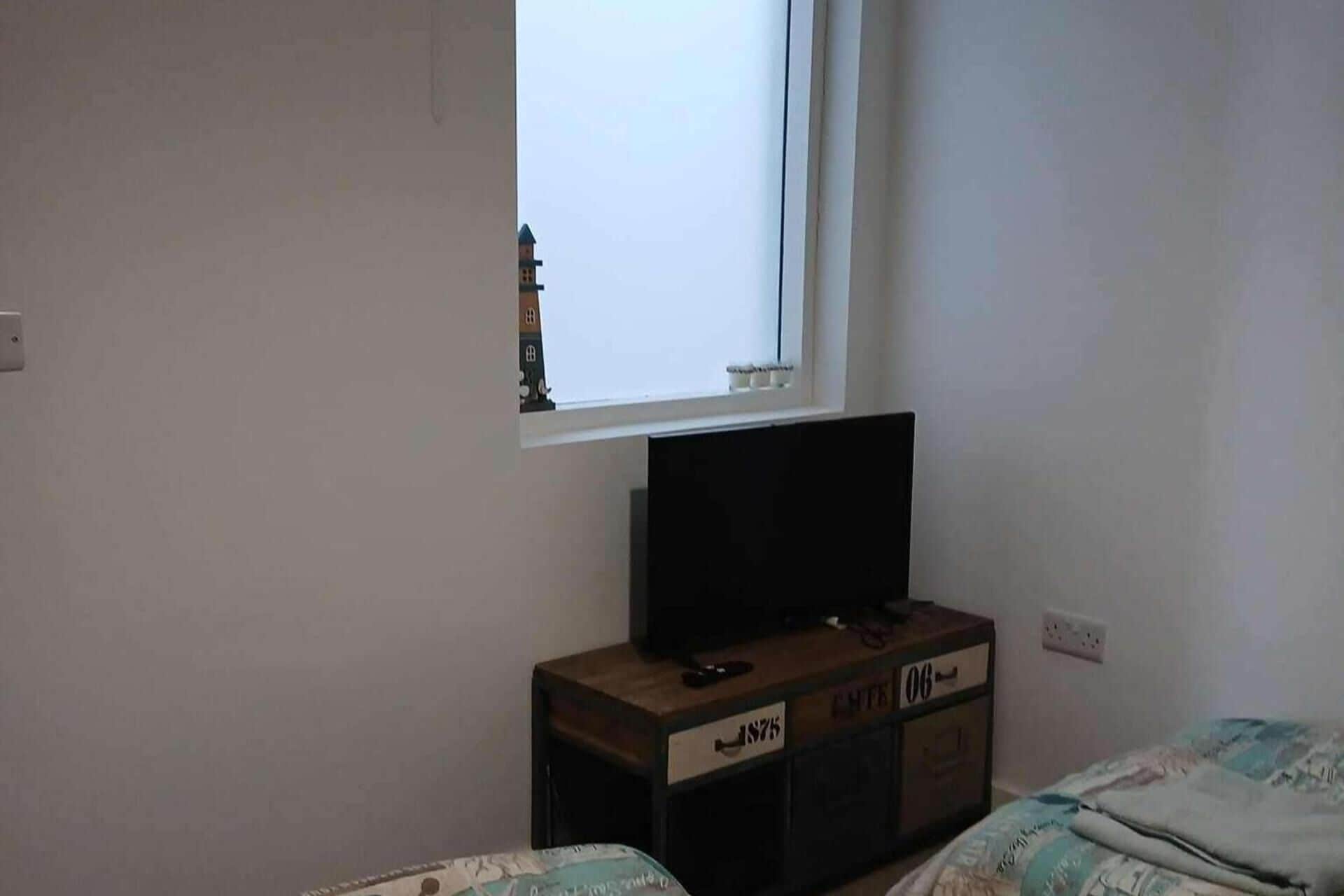 A bedroom with a single bed, a TV on a wooden dresser, and a window with a view of a building.