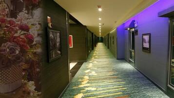 Un couloir d’hôtel avec un sol recouvert de moquette, des tableaux encadrés accrochés aux murs et un plafond éclairé d’une lumière violette.