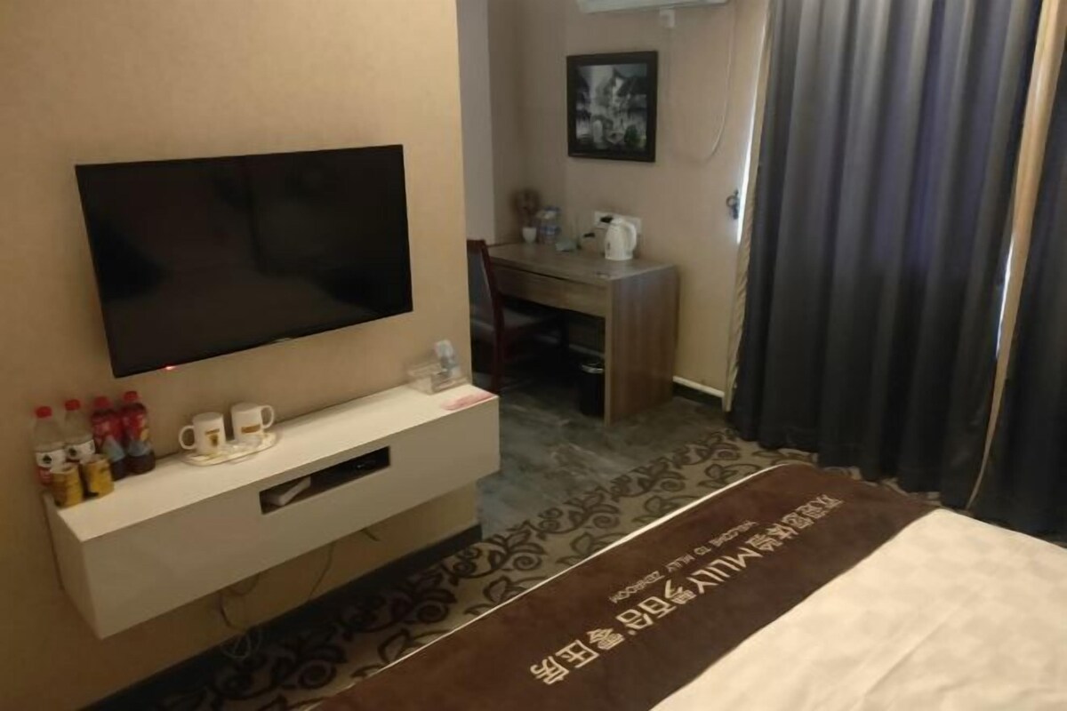 Ein Hotelzimmer mit Flachbildfernseher auf einem Standfuß, einem Schreibtisch mit Stuhl und einem Bett mit dekorativer Kopfteil.