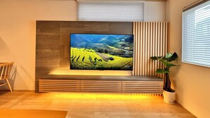A modern living room with a flat-screen TV mounted on a wooden panel wall, a potted plant, and a window with blinds. - AOI SUITE TAKAYAMA (Takayama)
