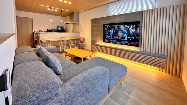 A modern living room with a grey sofa, wooden dining table, and a TV mounted on the wall. - AOI SUITE TAKAYAMA (Takayama)
