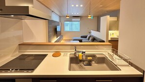 A modern kitchen with a built-in stove, sink, and a small island. There is a window with blinds, a sofa, and a dining area visible in the background. - AOI SUITE TAKAYAMA (Takayama)