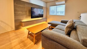 A modern living room with a flat-screen TV mounted on a wooden wall, a sofa, a coffee table, and a window with blinds. - AOI SUITE TAKAYAMA (Takayama)