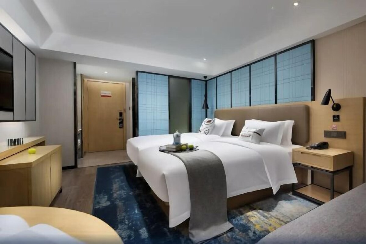 A modern hotel room with a large bed, bedside tables, a desk, and a large window with blinds.