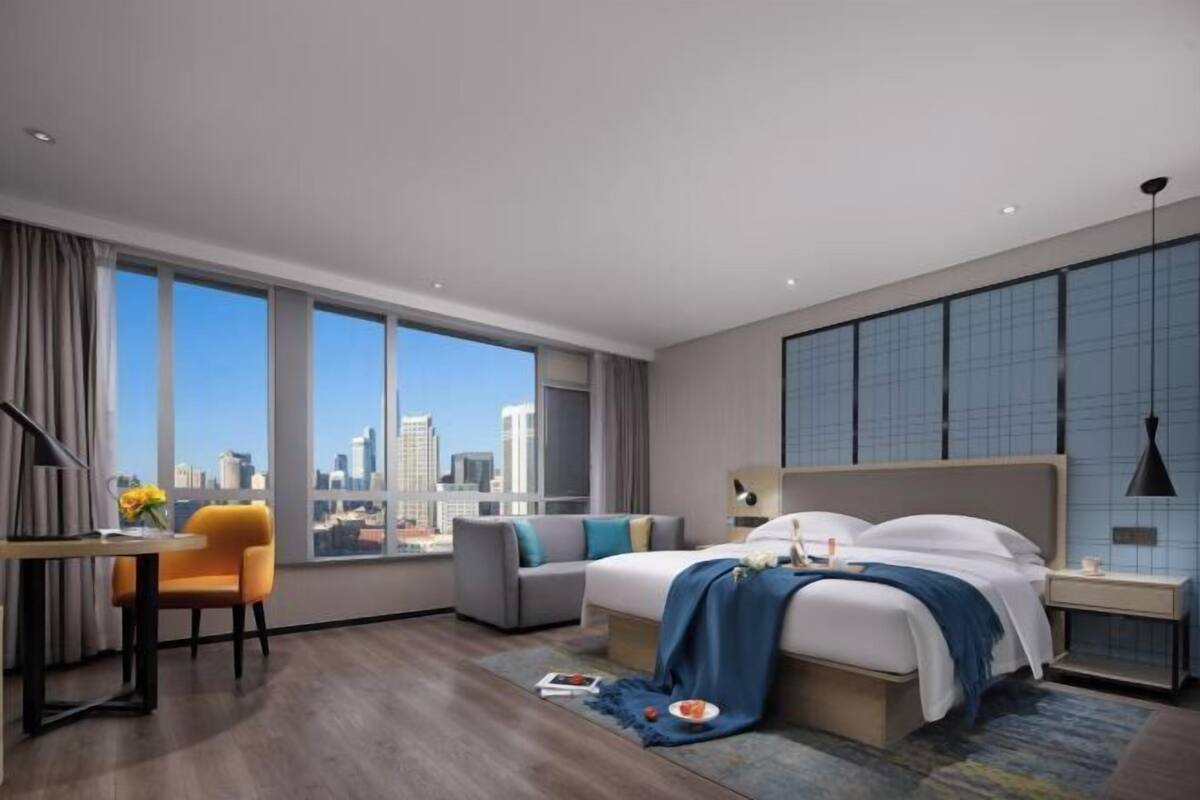 A modern hotel room with a large window, a bed, a desk, and a sofa.