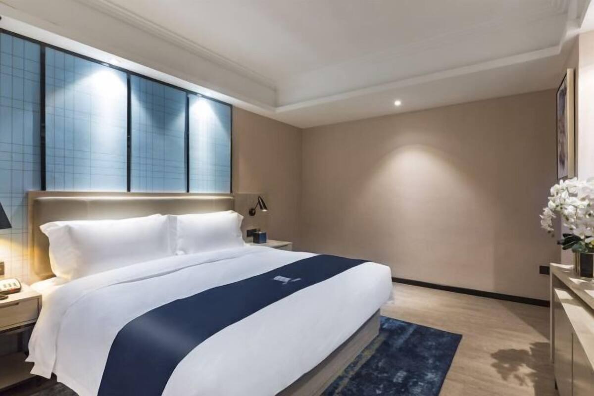 A hotel room with a large bed, a nightstand, a lamp, a vase with flowers, and a large window with blinds.