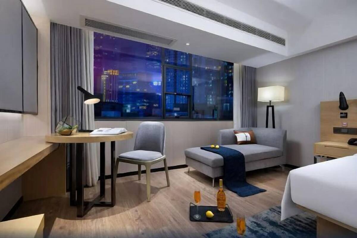 A hotel room with a large window, a desk with a lamp, a sofa, a bed, and a small table with a bottle and glasses.