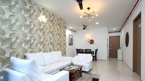 A modern living room with a white sofa, a coffee table, and a patterned wall. There is a dining area with a table and chairs in the background. - 3 BHK Urban Suite ( Gagal Home) (Mumbai)
