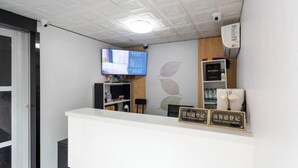 Zona de recepción con un mostrador blanco, un televisor montado en la pared y un estante con un jarrón y un letrero.
