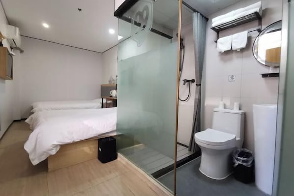 A modern hotel room with a bed, a glass shower enclosure, a toilet, and a small bedside table with a lamp.