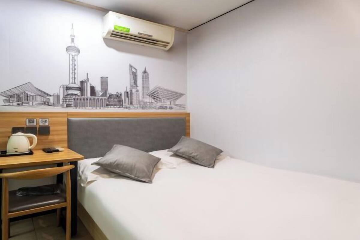 A hotel room with a bed, a desk with a kettle, a phone, and a wall mural of a city skyline.