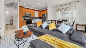 A modern living room with a grey sectional sofa, a wooden coffee table, and a dining area with a table and chairs. - 2335 Amazing 5BD Family Townhome Close to Disney (Kissimmee)
