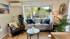 Una sala de estar acogedora con un sofá beige, una mesa de centro redonda y una mesa de comedor de madera. Hay un ventanal con vistas a un jardín y una valla de madera.