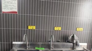 Un baño público con tres lavamanos, cada uno con un grifo y un dispensador de jabón verde. Hay letreros sobre los lavamanos que indican la disponibilidad de agua y jabón.