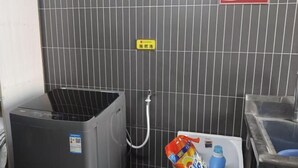 Un baño con pared de azulejos grises, un cubo de basura, un dispensador de agua y un inodoro con una botella de jabón para manos y una botella azul.