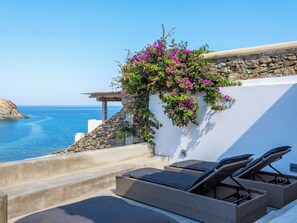 A seaside balcony with wicker lounge chairs, a stone wall, and a view of the sea. - Mykonist Merchia Beach Villas & Suites (Mykonos)