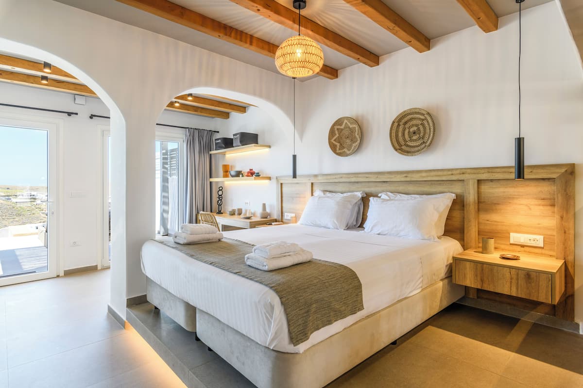 Un dormitorio moderno con una cama grande, cabecera de madera, mesitas de noche, una lámpara colgante y vistas a través de un puerta abierta.