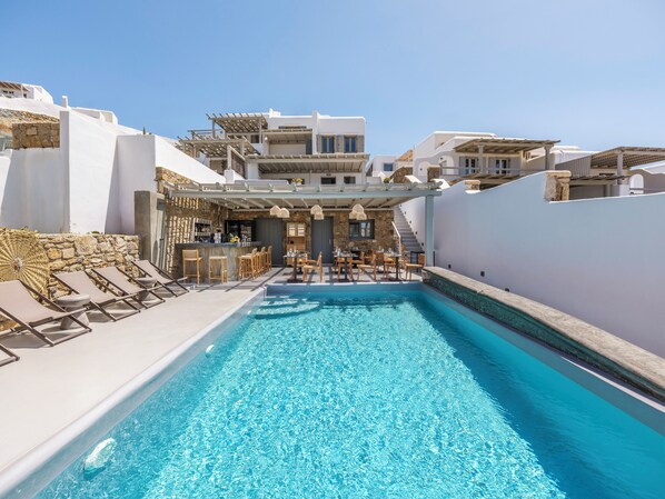 A swimming pool with lounge chairs and a bar area, surrounded by white buildings with stone accents. - Mykonist Merchia Beach Villas & Suites (Mykonos)