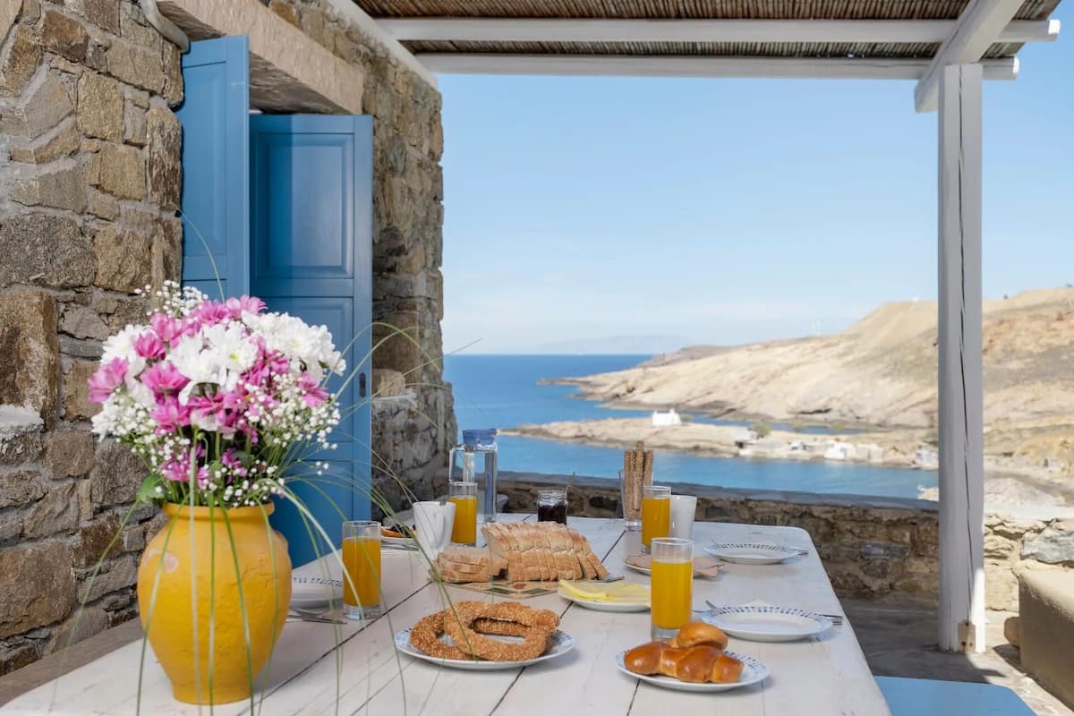 Mesa servida con alimentos para el desayuno y un jarrón con flores, con vista a un paisaje costero.