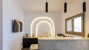 A modern bathroom with a curved mirror, a wall-mounted TV, and a countertop with a soap dispenser. - Mykonist Merchia Beach Villas & Suites (Mykonos)