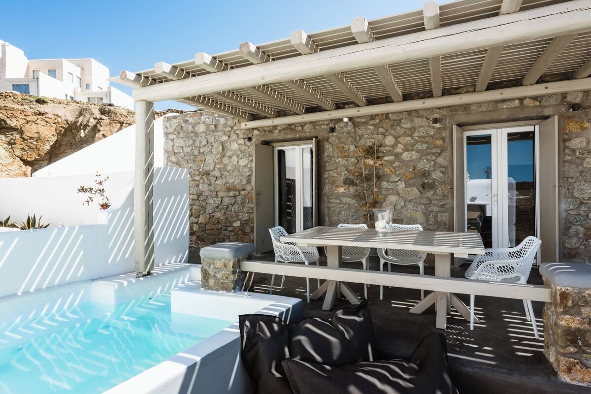 Zona de piscina con pared de piedra, mobiliario blanco y una pérgola.