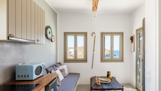 A compact living space with a kitchenette, a sofa, a dining table, and two windows offering views of the landscape. - Mykonist Merchia Beach Villas & Suites (Mykonos)