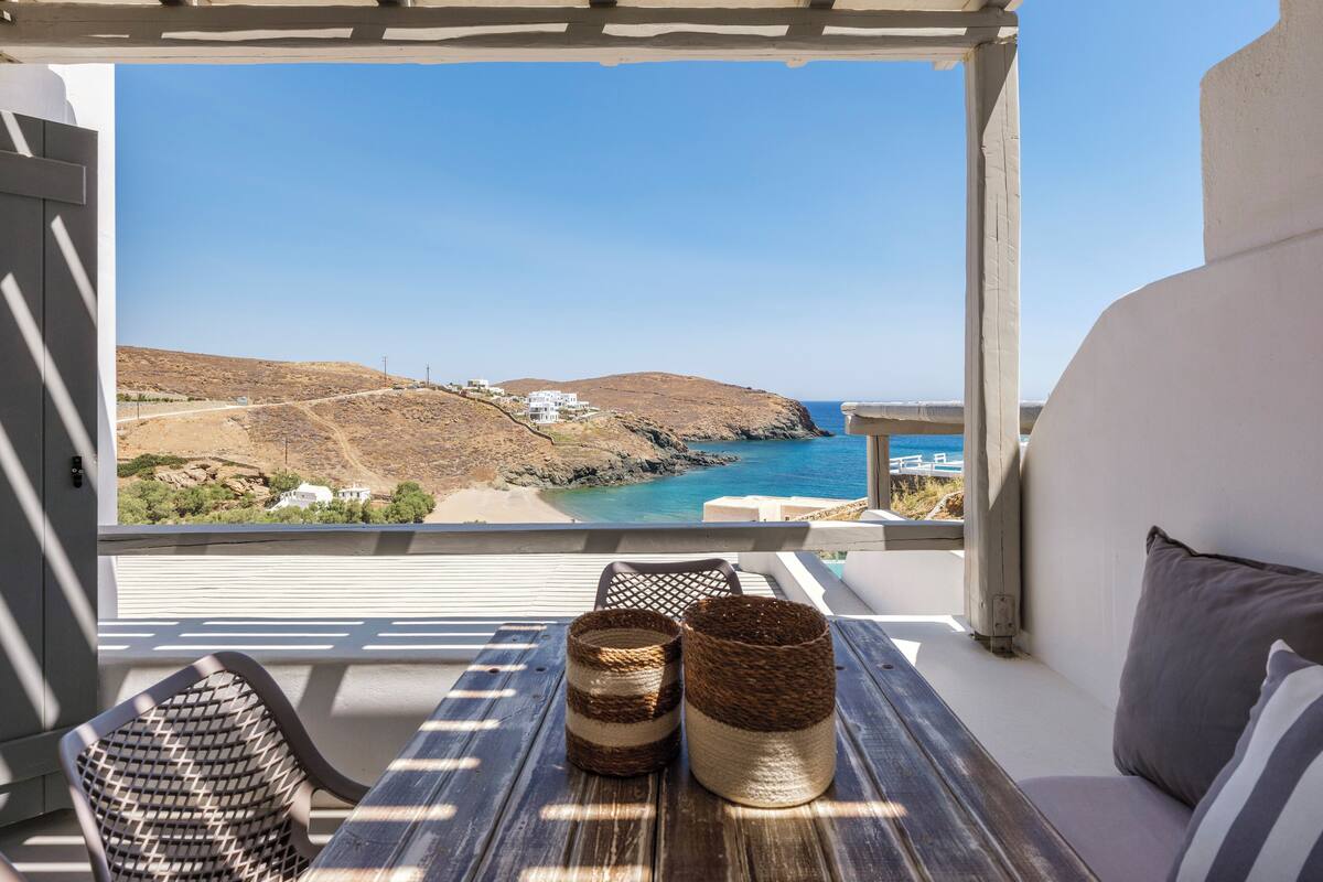 Un balcón con vistas al paisaje costero, una mesa de madera con dos cestas tejidas y un banco blanco acolchado.
