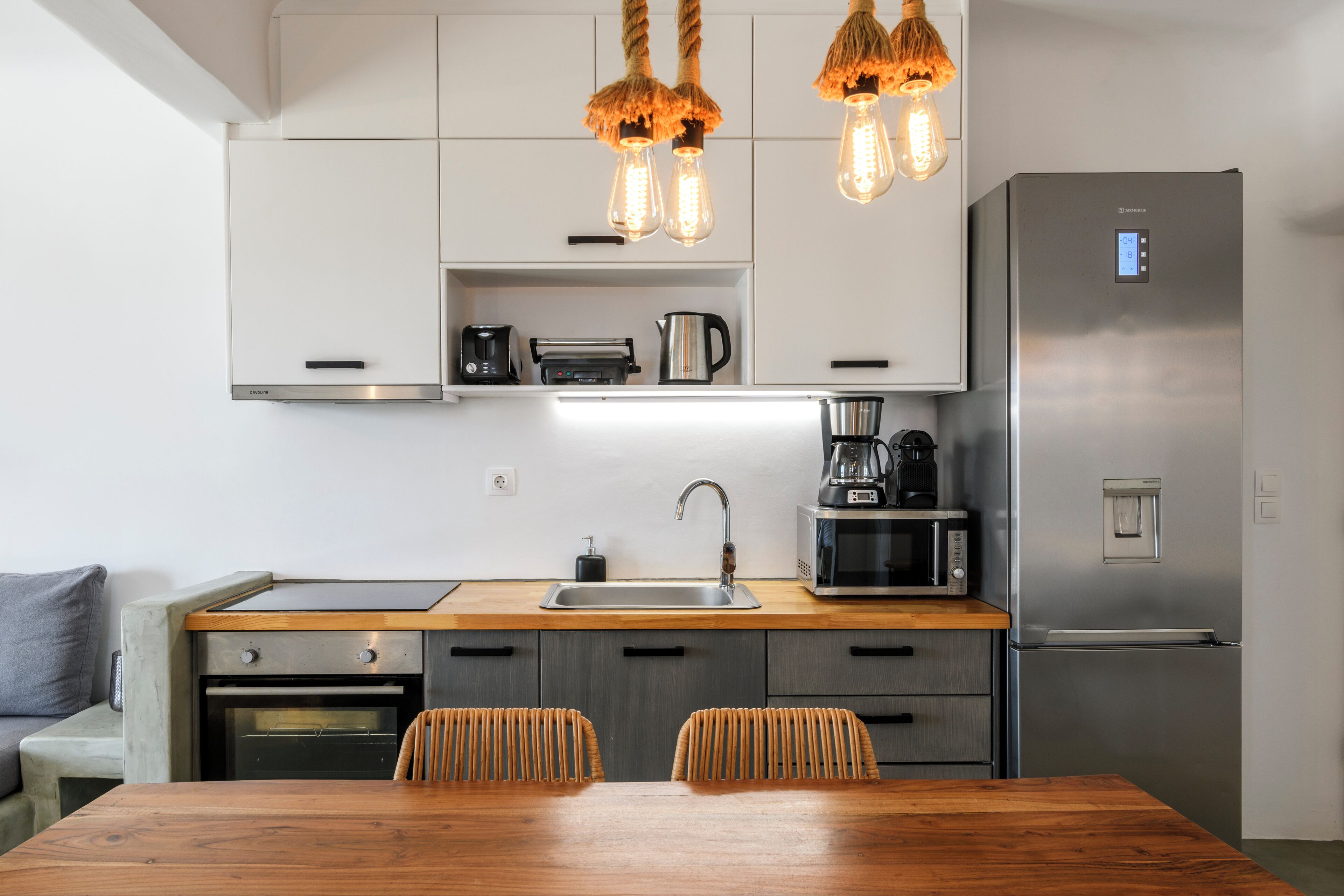 Una cocina moderna con un refrigerador de acero inoxidable, un horno empotrado, un microondas y una cafetera. Hay una mesa de comedor de madera con dos sillas.
