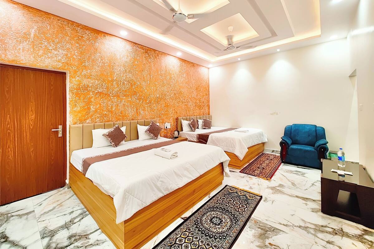 Quarto de hotel com duas camas, uma poltrona azul, uma porta de madeira e um tapete estampado.