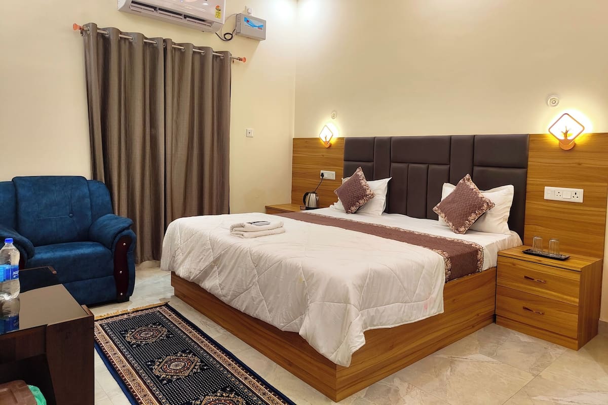Quarto de hotel bem arrumado, com cama, mesas de cabeceira, uma poltrona azul, uma janela grande com cortinas e um aparelho de ar condicionado.