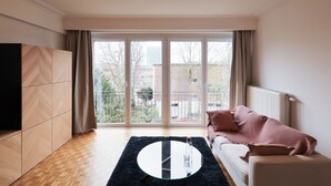 A modern living room with a large sliding glass door leading to a balcony, a white sofa with a pink throw, and a glass coffee table. - Fernand Cocq Elegant Residence (Brussels)