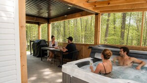 A covered outdoor area with a hot tub, a table with people dining, and a grill.