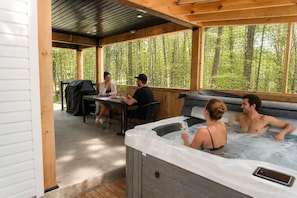 A covered outdoor area with a hot tub, a table with people dining, and a grill.