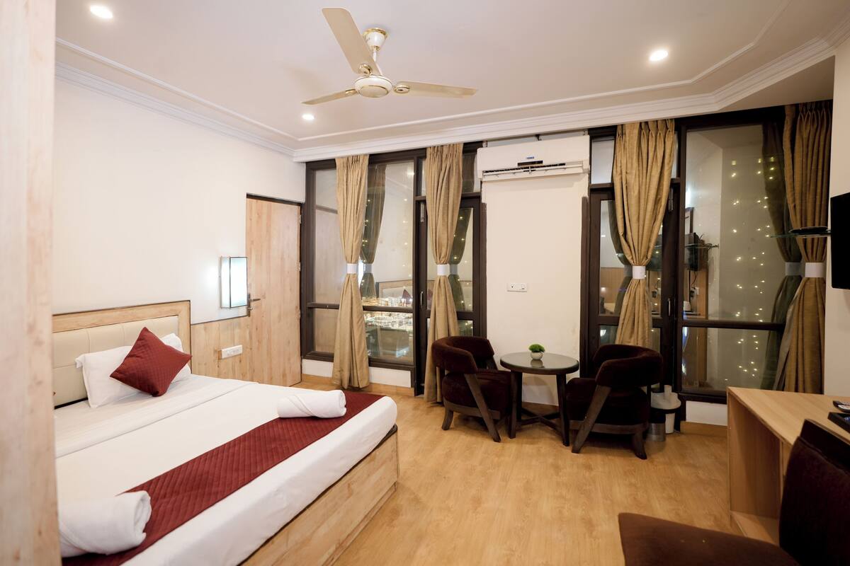 A hotel room with a bed, two chairs, a small table, a ceiling fan, and large windows with curtains.