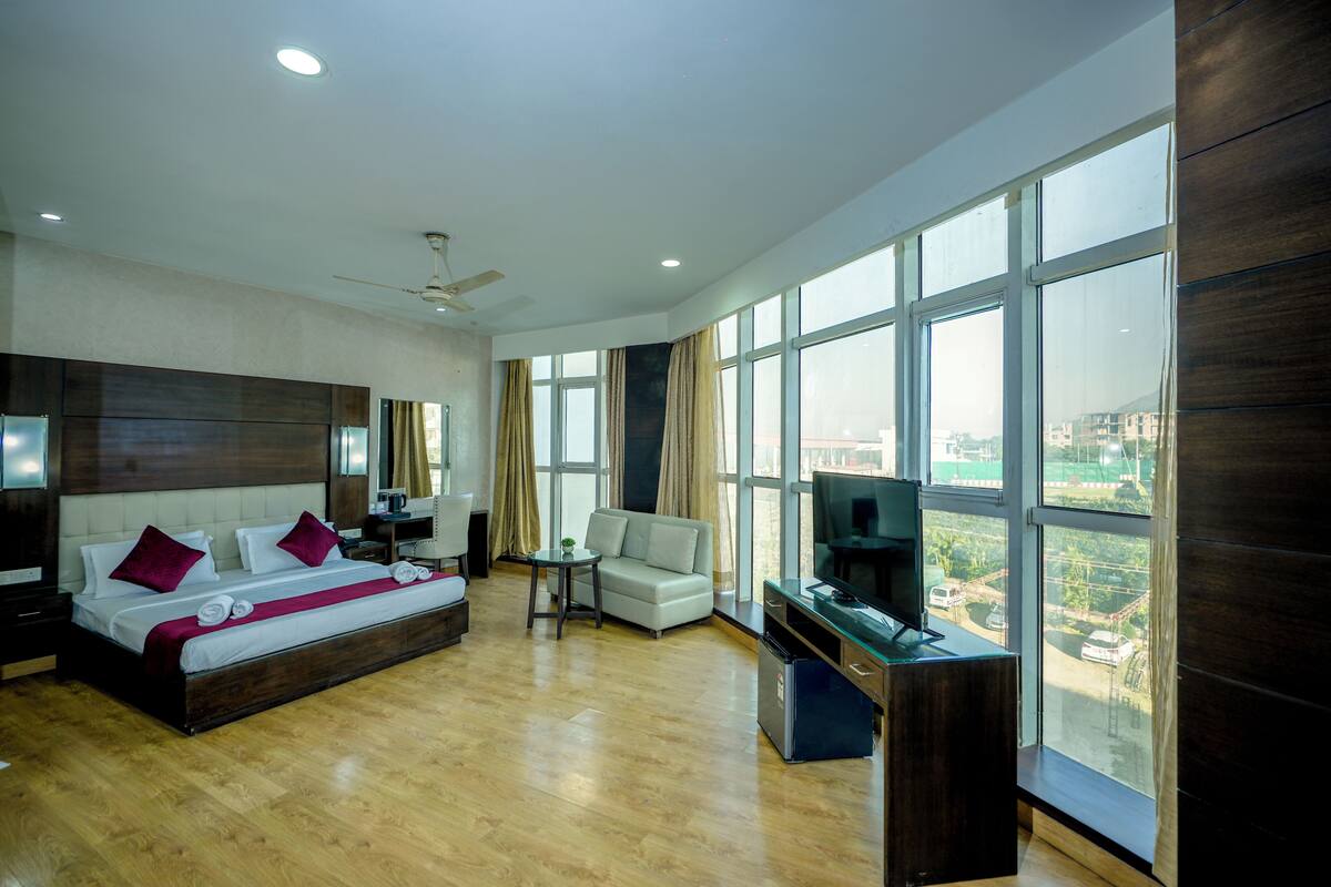 A spacious hotel room with a large bed, a sofa, a TV, and a view of the city through large windows.