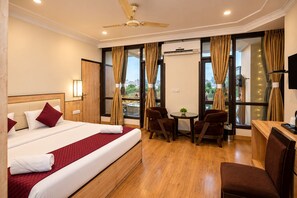 A hotel room with a bed, a desk, two chairs, a table, a ceiling fan, and large windows with curtains. - The Altruist Katra Hotel (Katra)