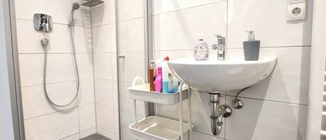 Un baño moderno con ducha a ras de suelo, un lavabo blanco con espejo y un estante pequeño con artículos de aseo.