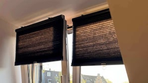 A window with two black blinds, a white window frame, and a view of buildings and trees outside. - Big Premium Apartment with Rhine View, totally new (Duisburg)