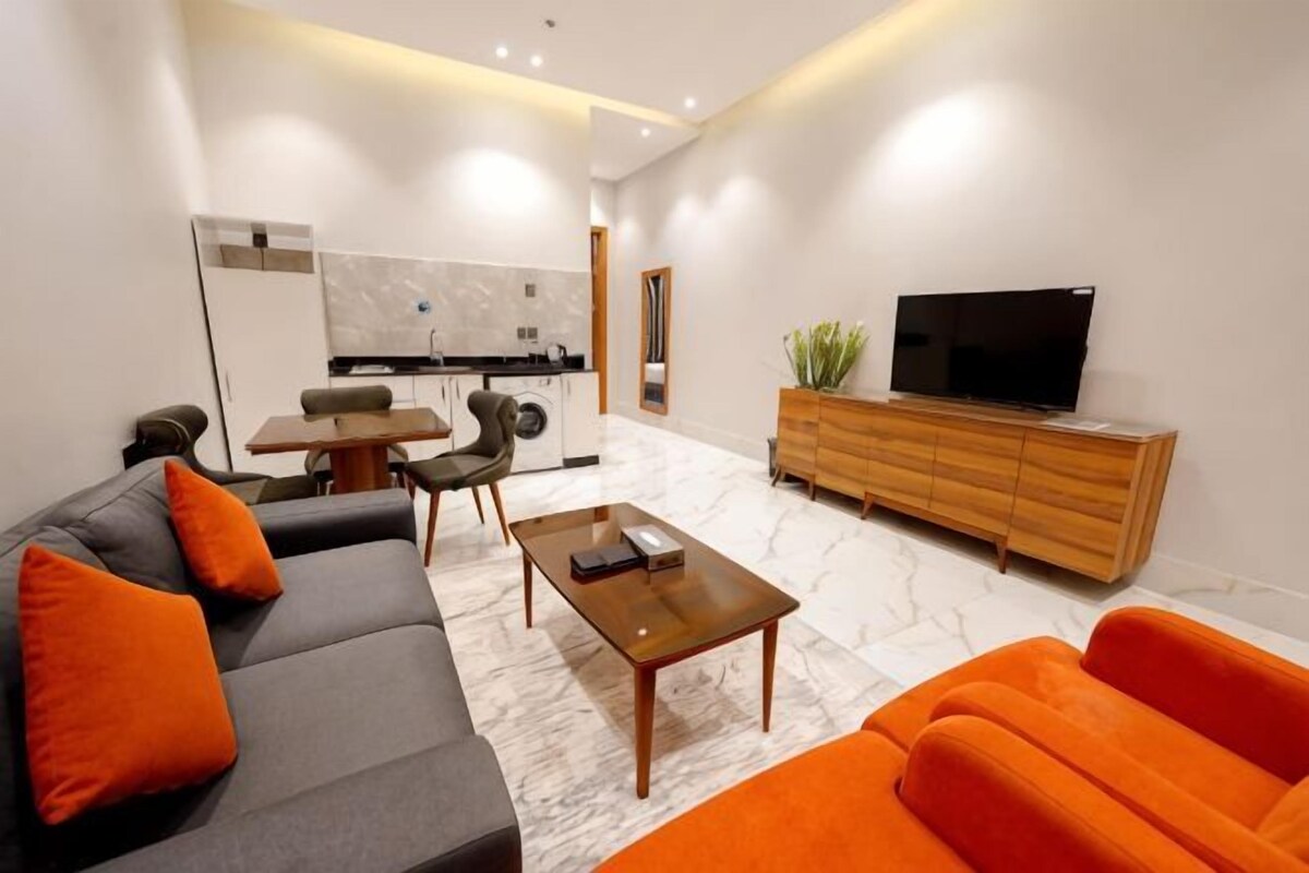 A modern living room with a sofa, coffee table, and TV, a dining area with chairs, and a kitchen in the background.