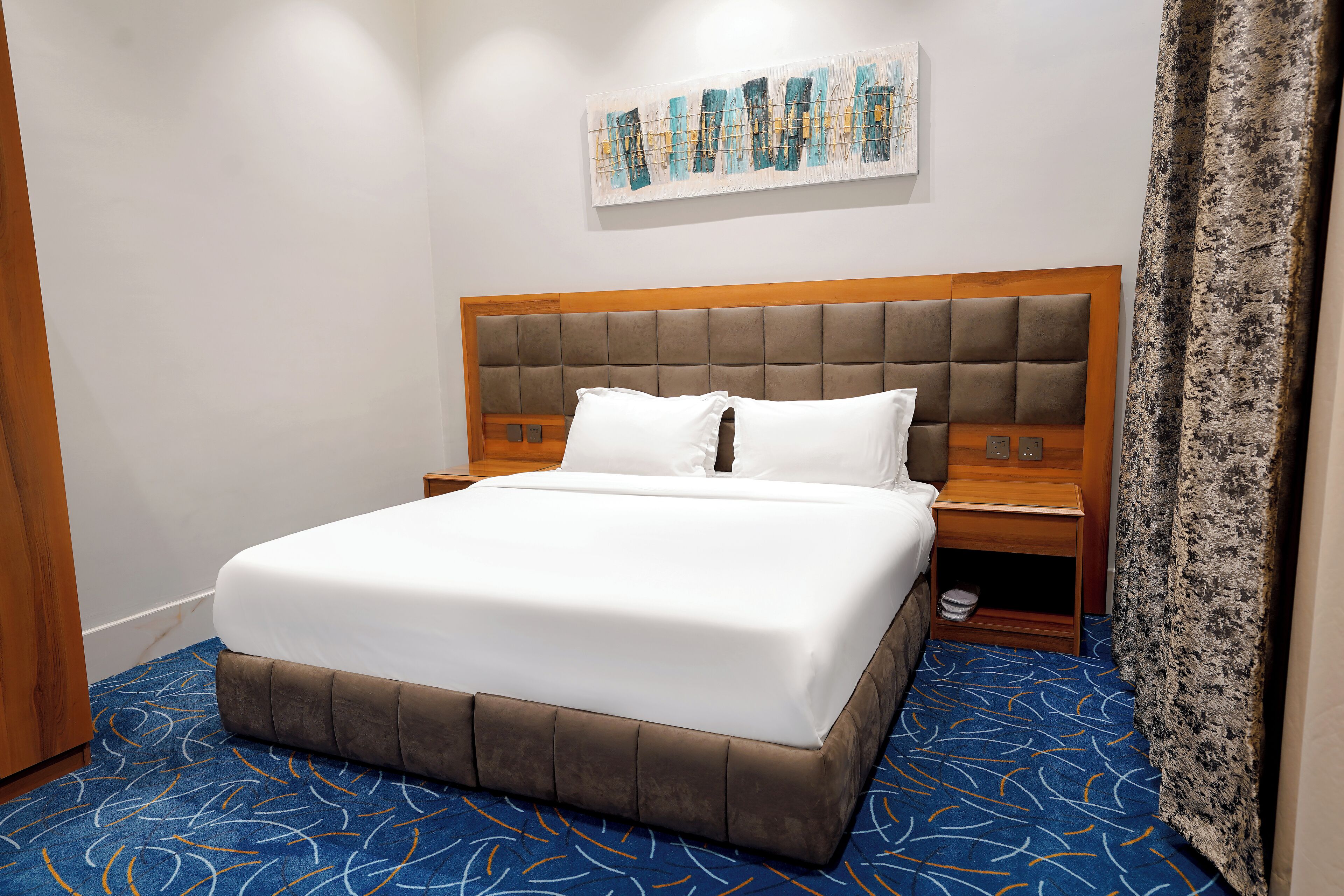 A hotel room with a large bed, a headboard with a padded design, a wooden nightstand, and a blue patterned carpet.