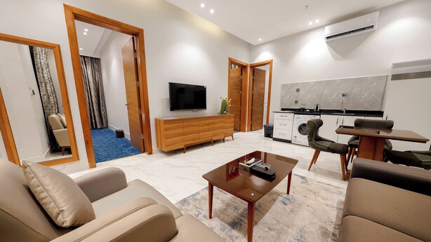 A modern living room with a sofa, coffee table, and a TV. There is a kitchen area with a dining table and chairs.
