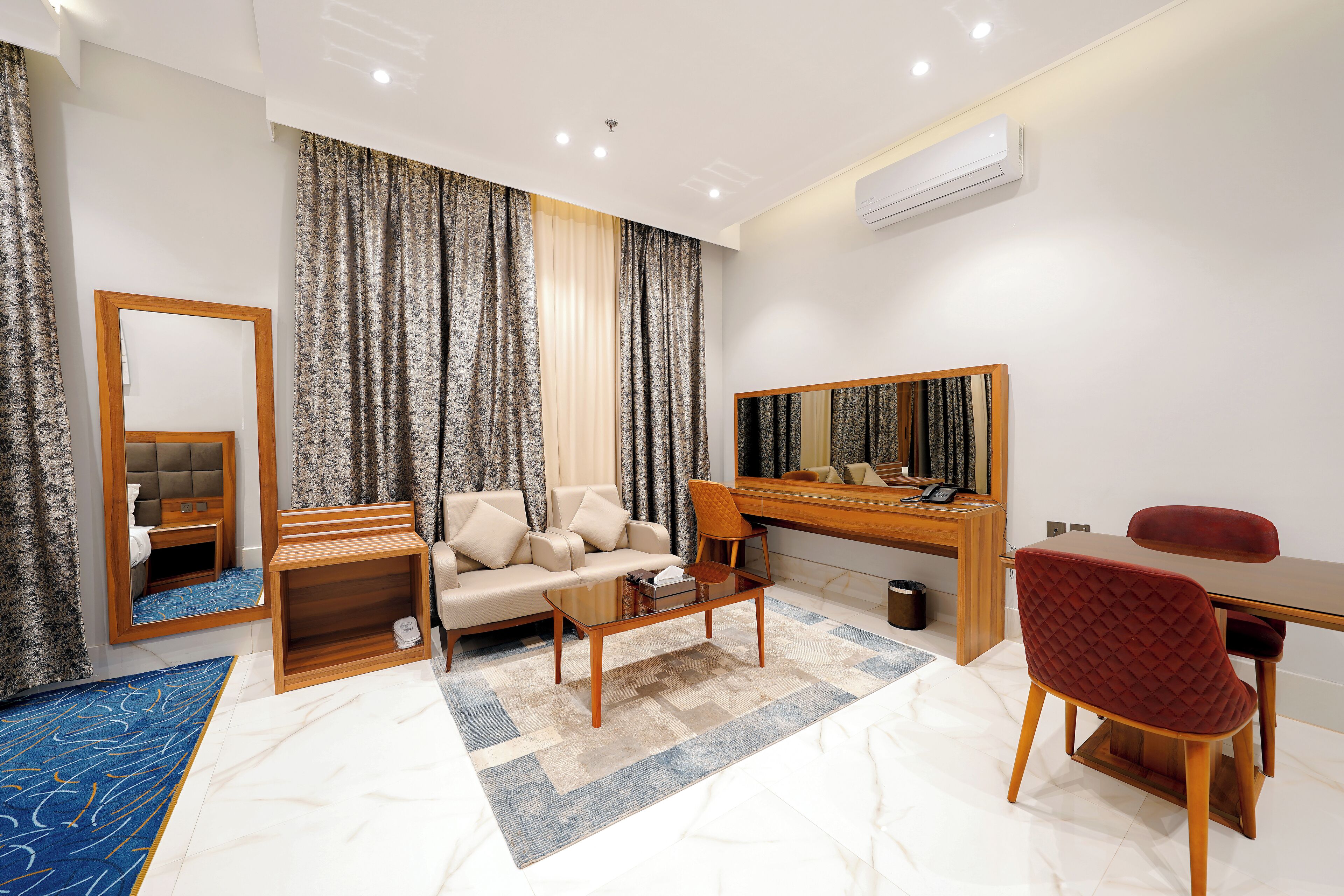 A modern hotel room with a sofa, a desk, a chair, a mirror, and a small seating area.