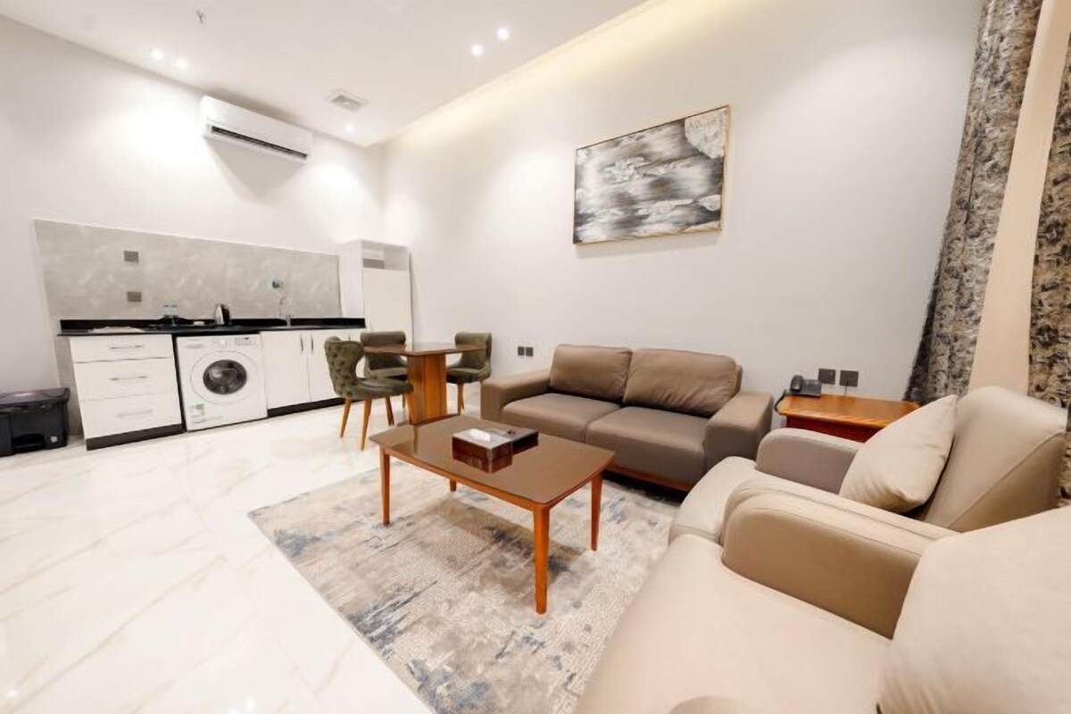 A modern living room with a sofa, coffee table, and chairs. A kitchen with a washing machine and cabinets is visible in the background.