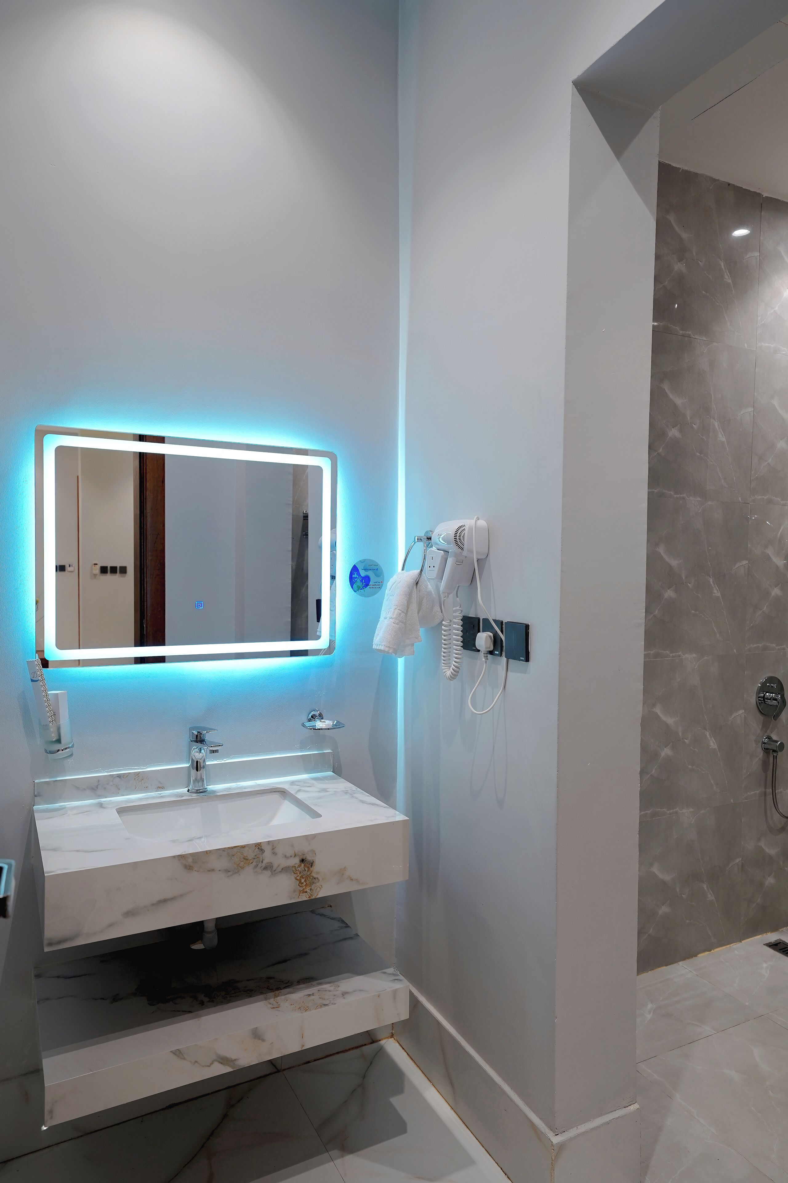 A modern bathroom with a marble sink, a mirror with LED lighting, a hairdryer, and a shower area.