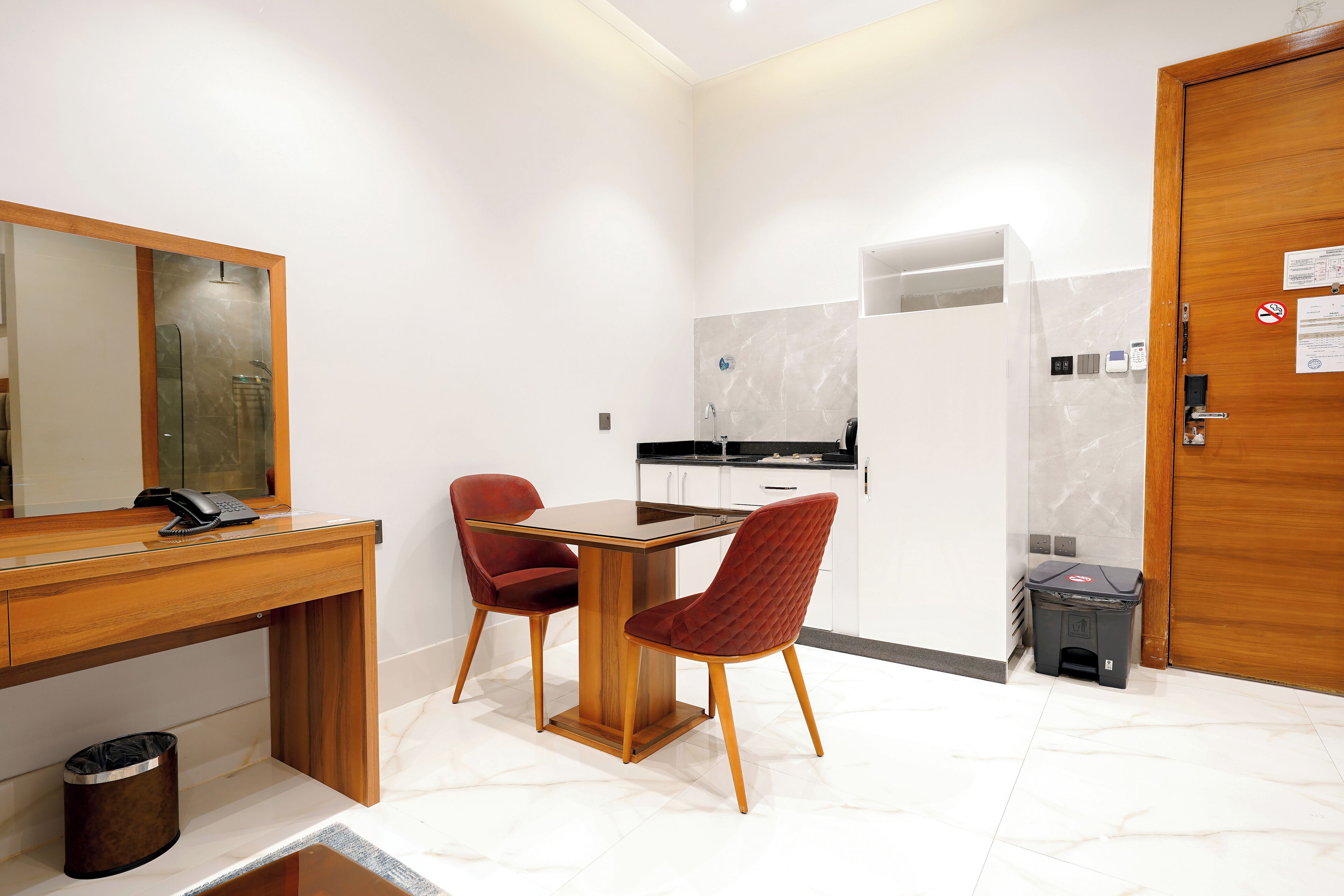 Une chambre d’hôtel compacte avec un bureau en bois, deux chaises rouges, une petite poubelle et une porte en bois.
