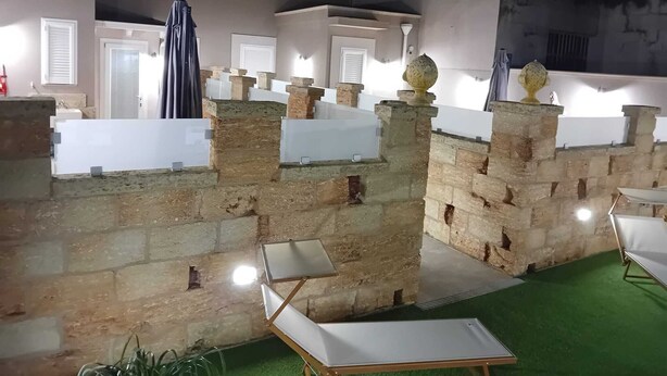 Ein Innenhof mit Steinmauern, weißen Säulen und einem grünen Rasen. Zwei weiße Liegestühle und ein kleiner Tisch sind ebenfalls vorhanden.