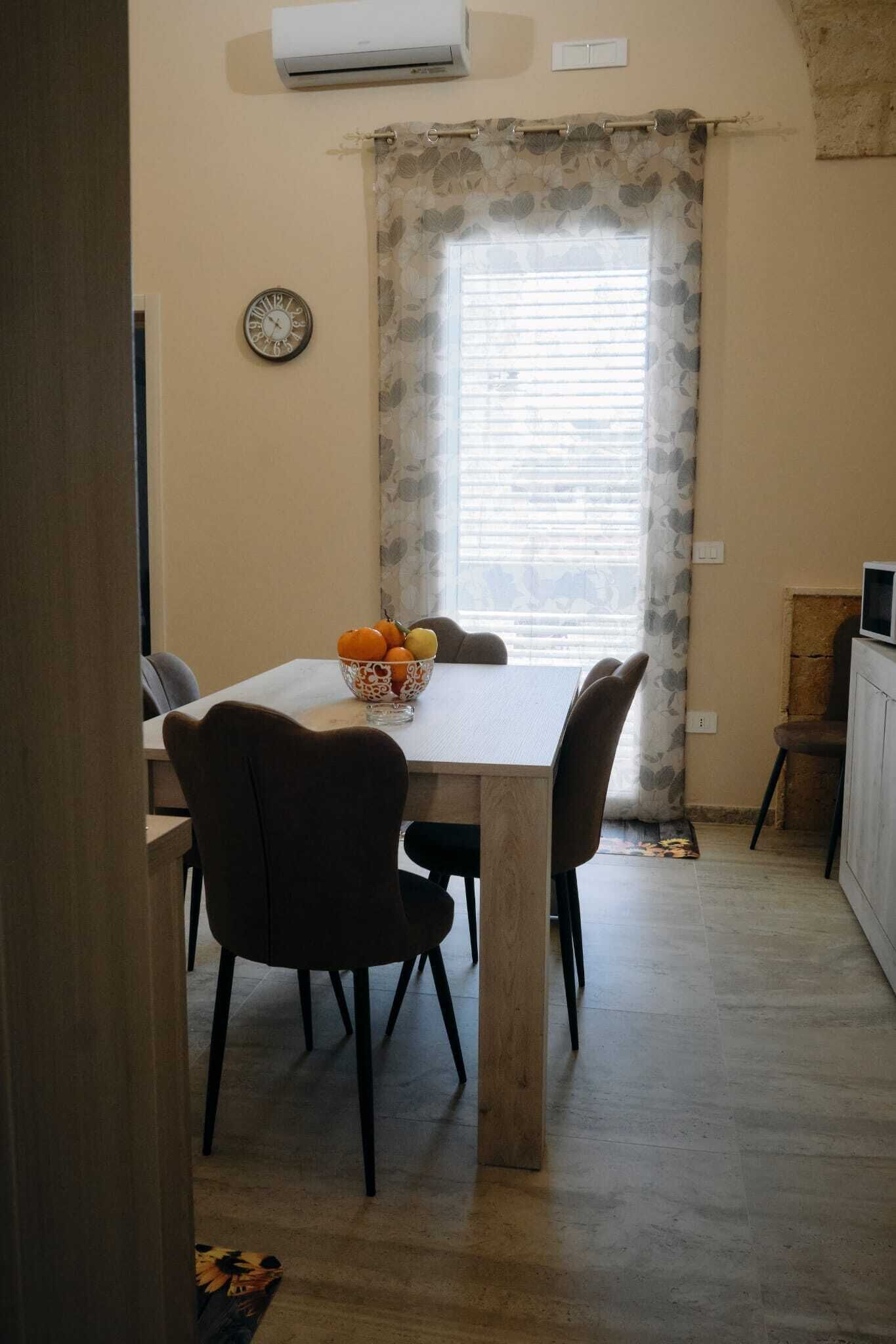 Un área de comedor con una mesa, sillas, un cuenco de fruta, un reloj y una ventana con persianas.