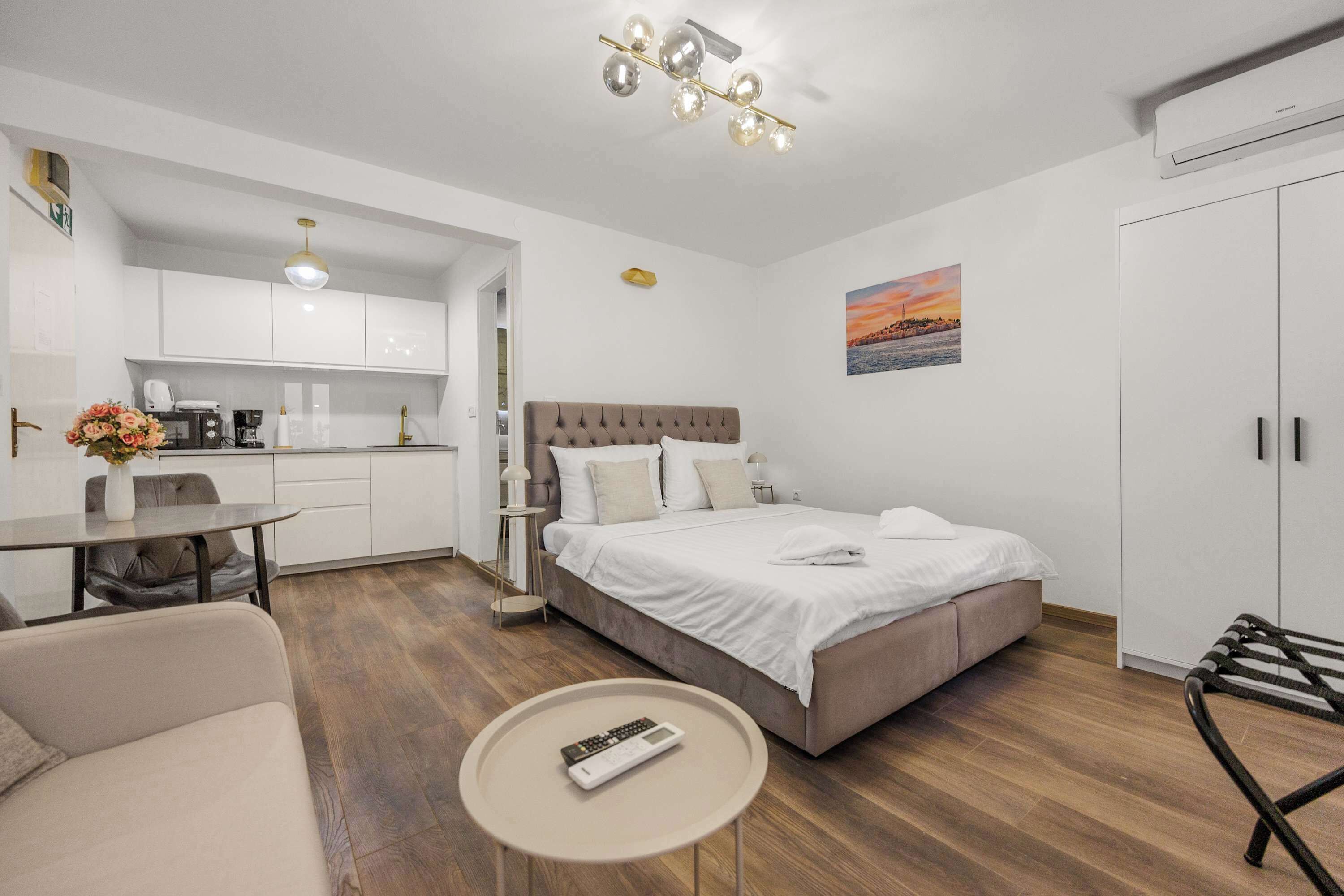 A modern hotel room with a bed, a small round table, a sofa, a kitchenette, and a view of the sunset.