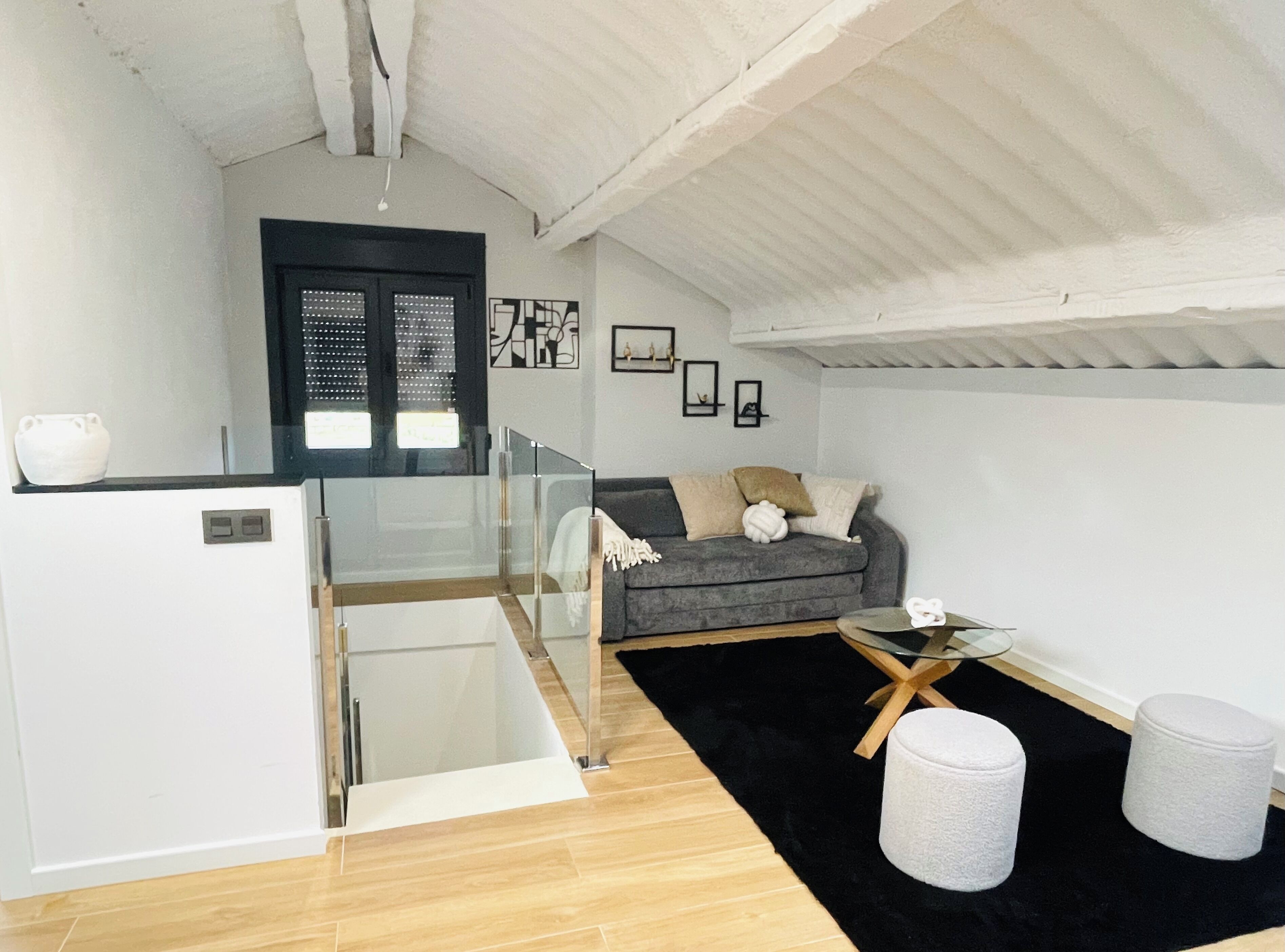 A modern living room with a grey sofa, a small round table, and a white ottoman. There is a black and white rug, a white cabinet, and a black-framed window.
